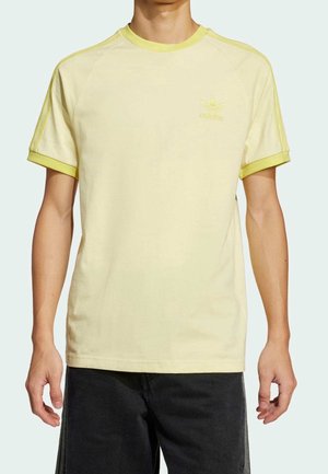 Mężczyzna w jasnożółtej koszulce Adidas z krótkim rękawem i trzema paskami na rękawach oraz czarnych szortach, stojący na jednolitym tle.