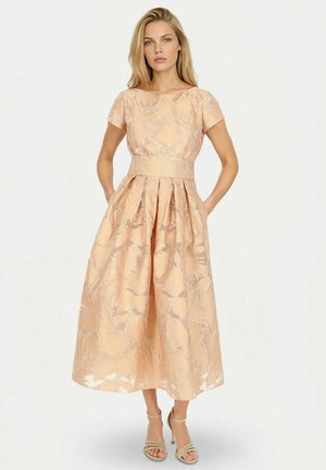 Kobieta o blond włosach, ubrana w midi sukienkę w kolorze brzoskwiniowym z kwiatowym wzorem, krótki rękaw, beżowe sandały na obcasie, stoi z rękami w kieszeniach.