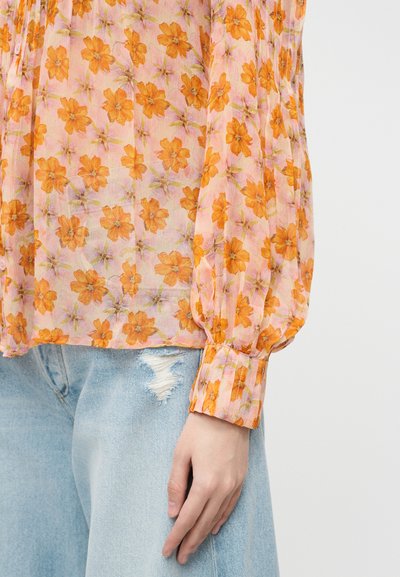 Διάφανη μπλούζα με πορτοκαλί λουλουδάτο σχέδιο και μακριά μανίκια με μανσέτες, συνδυασμένη με ανοιχτό μπλε τζιν με σκισίματα.
