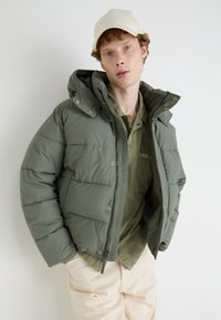 Green puffer jacket with an adjustable hood, zip closure, and snap buttons. Paired with a light green shirt and beige pants, standing against a plain background.