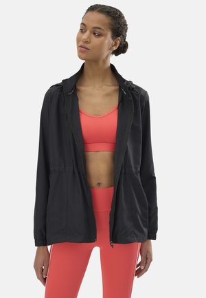 Mujer con sujetador deportivo coral y leggings, con chaqueta negra con cremallera, de pie y mirando ligeramente de lado contra un fondo liso.