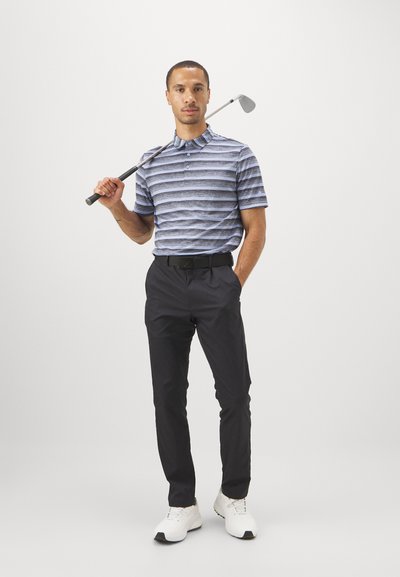 Golfa polo krekls ar zilu un pelēku svītru rakstu, melni bikses, tur rokā golfu nūju. Balti apavi pabeidz sportisko apģērbu.