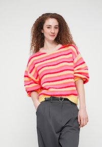 Γυναίκα με κατσαρά κόκκινα μαλλιά που φοράει πολύχρωμο ριγέ πουλόβερ, γκρι παντελόνι και μαύρη ζώνη, στέκεται με το ένα χέρι στην τσέπη.