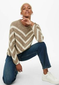 Pull à motif chevron beige et blanc avec un col rond, associé à un jean bleu et des baskets blanches. La texture semble douce et tricotée.