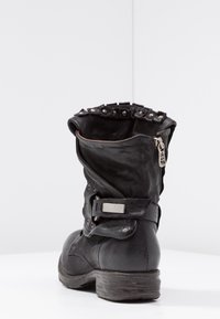 Bota de tobillo de cuero negro con parte superior doblada, tachuelas plateadas alrededor del borde, cremallera lateral y tacón bajo apilado. Material texturizado y diseño robusto.