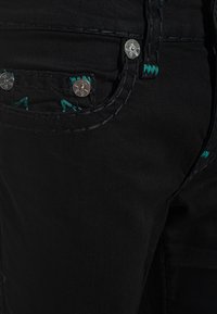 Plano detalle de un bolsillo de vaqueros de mezclilla negra con remaches plateados y detalles de costuras en color verde azulado en las costuras y la presilla del cinturón.