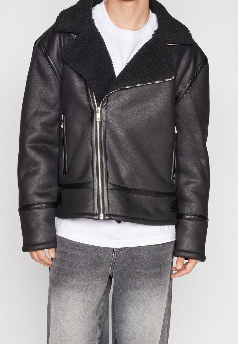 Sort jakke i kunstlæder med krave i imiteret pels, lynlås foran og brede ærmer. Har to sidelommer og kontrasterende sorte detaljer.