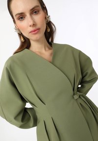 Olive green wrap dress with wide sleeves, textured fabric, and a belted waist. Features gold and floral statement earrings.