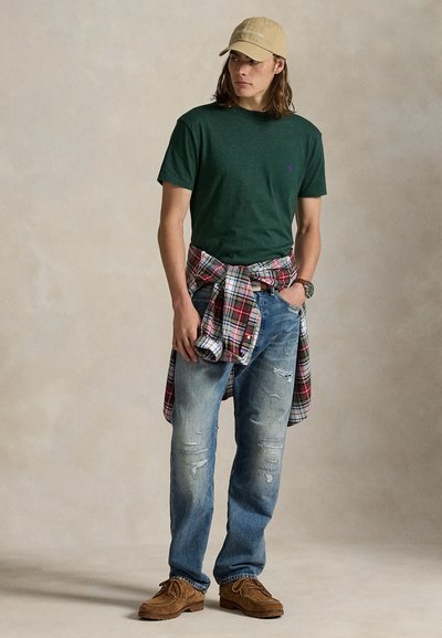 Πράσινο t-shirt με στρογγυλή λαιμόκοψη, μπλε τζιν με φθορές, κόκκινο καρό πουκάμισο φλατλάνελ δεμένο γύρω από τη μέση, μπεζ καπέλο, καφέ παπούτσια. Απλή καθημερινή εμφάνιση.