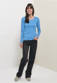 Long-sleeve light blue shirt paired with black pants. Model stands on a neutral background, wearing green and white sneakers.