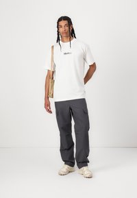 Camiseta blanca con logo negro, pantalones cargo grises y zapatos beige; modelo sosteniendo un bolso de hombro beige. Diseño minimalista, ajuste casual.