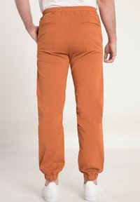 Orange joggers med elastisk midja och muddar, med två bakfickor och en slät bomullstextur. Bärs med vita sneakers.