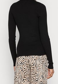 Svart, rutor med lång ärm och figursydd siluett; ihop med mönstrade beige och svarta byxor med virvlande mönster.