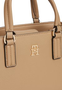 Μπεζ δερμάτινη τσάντα χειρός με διπλές λαβές και χρυσούς αρχικά λογότυπους "TH" στην υφή μπροστινή επιφάνεια.