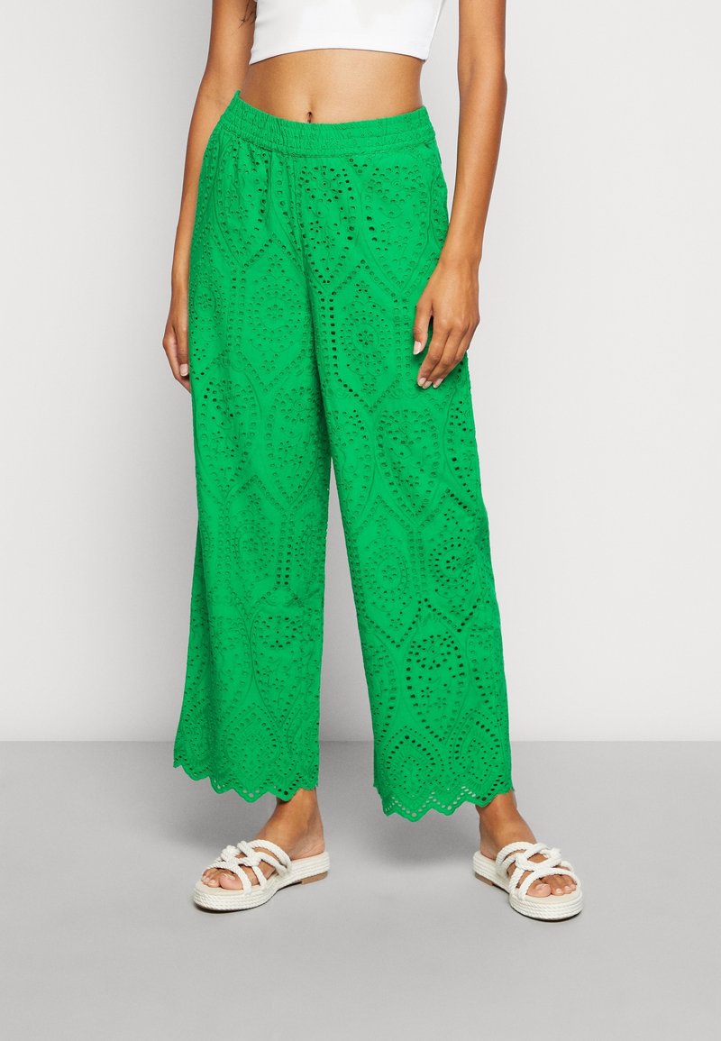 Pantalones verdes de pierna ancha con patrón de encaje de ojales y dobladillo en forma de ondas, usados con sandalias blancas y un top corto blanco, mostrados desde la cintura hacia abajo.