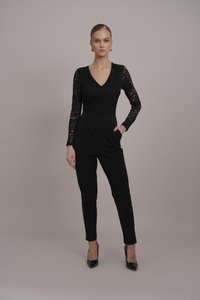 Combinaison noire avec des manches longues en dentelle, décolleté en V, jambes fuselées et poches latérales. Associée à des escarpins noirs à bout pointu.