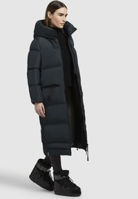 Giacca imbottita nera con colletto alto, silhouette lunga e tasche con zip. Il modello indossa stivali neri con lacci e suola spessa.