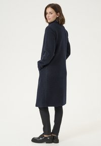 Marinblå texturerad lång kappa med sidofickor, hög krage och avslappnad passform, kombinerad med åtsittande svarta byxor och loafers.