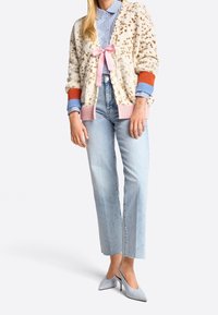 Gestructureerd crèmekleurig vest met bruine vlekken, roze en oranje accenten, gedragen over een blauw-wit gestreept shirt, gecombineerd met lichte spijkerbroek en grijze hakken.