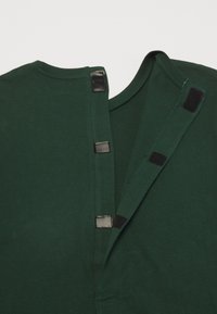 Zelený bavlněný oděv s kulatým výstřihem a patentkovým zapínáním, s černými akcenty podél otvoru a bočního švu.