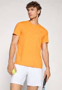 Oranž tenniseelik, millel on tekstuuriga triibuline muster, lühikesed varrukad ja ümmargune kaelus, koos valgete lühikesed pükstega ning hoiab tennisepalli.