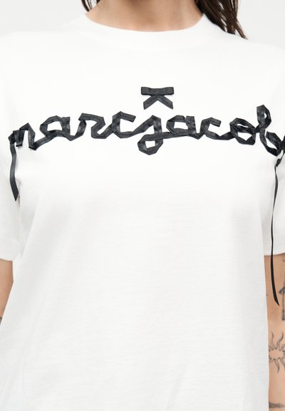 Λευκό μπλουζάκι με μαύρη κορδέλα που σχηματίζει χειρόγραφη γραφή στο στήθος και μικρό μαύρο φιόγκο πάνω από το κείμενο.