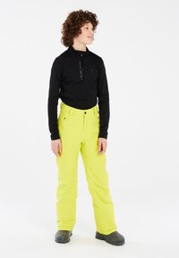 Zwarte longsleeve top met halve rits en logo, gecombineerd met felgele sneeuwbroeken met zakken, gedragen met zwarte waterdichte boots.