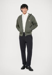 Hoodie zippé vert avec poches avant, arborant un petit logo coloré. Porté par-dessus une chemise blanche et associé à un pantalon et des chaussures sombres.