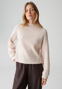 Maglione in maglia testurizzata rosa chiaro con collo alto, spalle scese e maniche lunghe, abbinato a pantaloni marrone scuro.