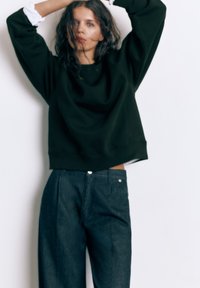 Žena valovite kose nosi crnu oversized majicu i tamne široke traperice, stoji s podignutim rukama ispred jednobojne pozadine.