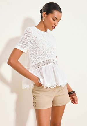 Femme aux cheveux en chignon portant un haut blanc en dentelle à manches courtes et un short beige avec des détails lacés sur les côtés, debout avec une main dans la poche.