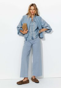 Chemise en denim bleu clair avec des coutures délicates, jeans évasés assortis et un gilet texturé bleu. Accompagnée de mocassins imprimés léopard.