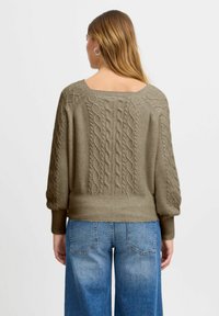 Olivgrüner Strickpullover mit weitem Ausschnitt und Ballonärmeln. Rippbündchen und Saum sorgen für Textur. Getragen mit hellblauen, weit geschnittenen Jeans.