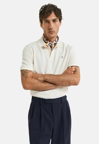 Cremfarbener Poloshirt mit Kragen, mit kleinem Logo; kombiniert mit dunkelblauen Hosen. Der Schal hat ein buntes, gemustertes Design.