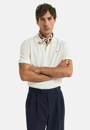 Polo shirt crema con colletto, dotata di un piccolo logo; abbinata a pantaloni blu scuro. La sciarpa presenta un design colorato e fantasioso.