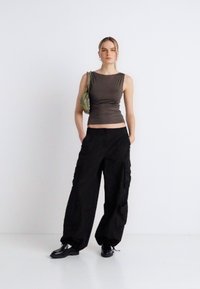 Haut gris à une épaule, coupe ajustée. Pantalon cargo noir avec poches latérales, coupe ample. Modèle tenant un petit sac vert, debout sur un fond blanc.