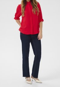 Blusa rossa con bottoni, maniche corte a volant, abbinata a jeans dritti scuri e décolleté crema a punta.