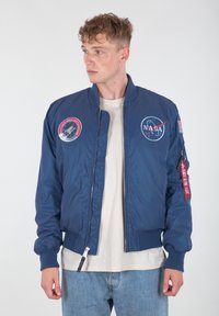 Granatowa kurtka bombowca z naszywkami NASA, ściągaczami przy mankietach i u dołu, zamek błyskawiczny oraz czerwoną metką na lewym rękawie. Gładki materiał nylonowy.
