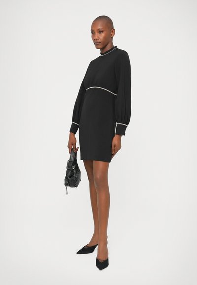 Vestido negro de manga larga con cuello alto, que presenta un ribete plateado en la cintura y los puños. Combinado con tacones negros puntiagudos y un bolso texturizado.