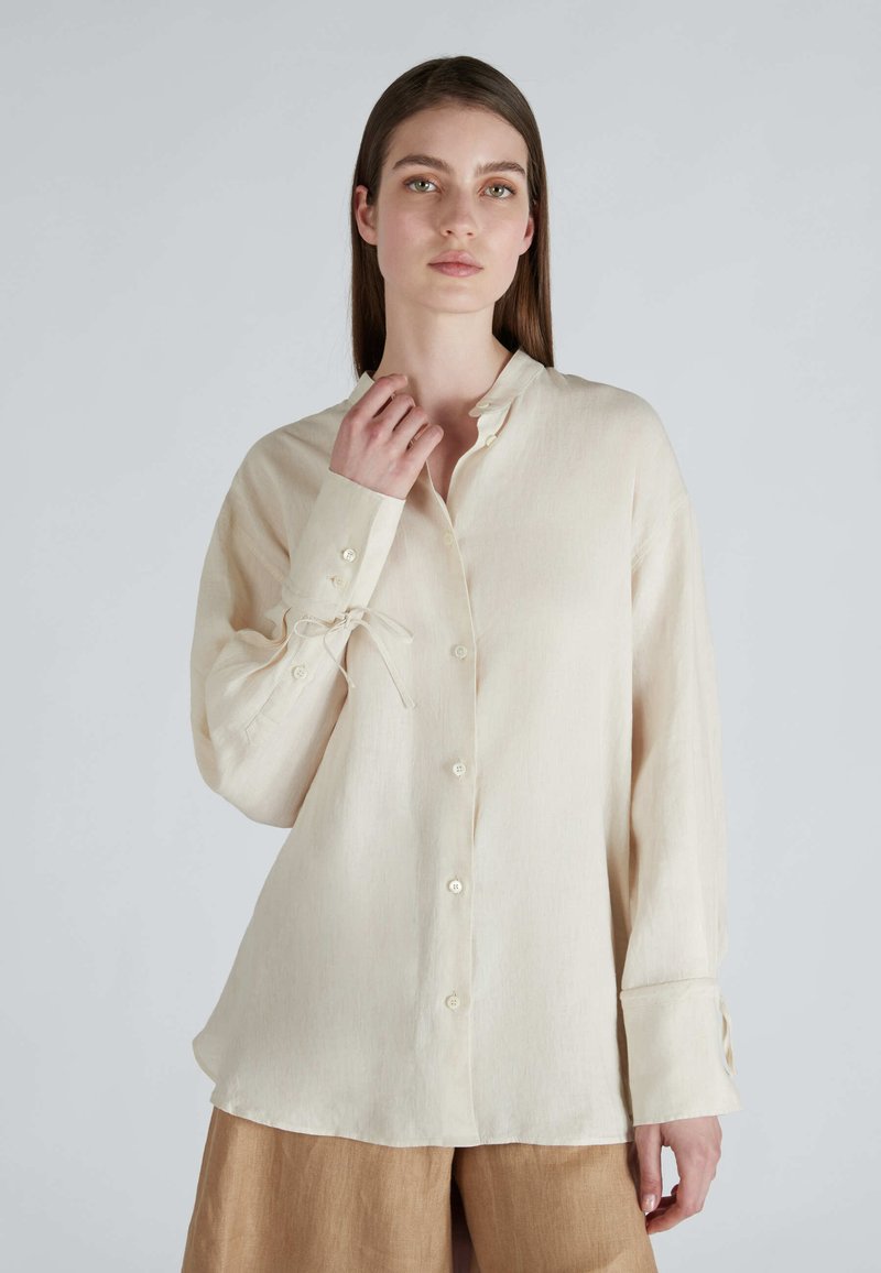 Camicetta a maniche lunghe color beige chiaro con colletto alla mandarino, apertura frontale con bottoni e dettagli con fiocco ai polsini, realizzata in un tessuto liscio e texturizzato.