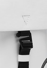 Svart justerbart band med plastspänne fäst vid en vit panel, som har en sydd triangulär design nära toppen.