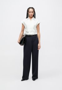 Λευκό μπλουζάκι με κοντά μανίκια και πιέτες, συνδυασμένο με ψηλόμεσο σκούρο μπλε παντελόνι. Το μοντέλο κρατά μαύρο φάκελο. Ομαλές υφές και εφαρμογή ραμμένη στα μέτρα.