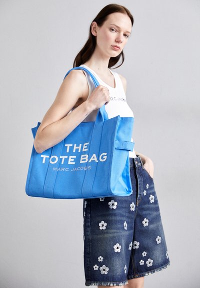 Borsa a trapezio blu in tela con testo bianco "THE TOTE BAG MARC JACOBS," dotata di due manici e forma strutturata, tenuta da un modello.