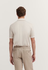 Beige Poloshirt mit kurzen Ärmeln und strukturiertem Fischgrätenmuster, kombiniert mit hellbeigen Hosen mit Gesäßtaschen und goldenen Akzenten.