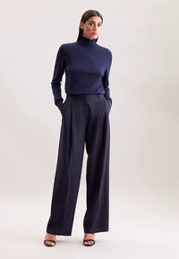 Marineblauwe coltrui gecombineerd met wijde marineblauwe broek. Soepele textuur, afgewerkte pasvorm, aangevuld met open-toe zwarte hakken. Eenvoudig ontwerp.