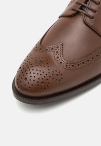 Scarpa da uomo in pelle marrone con design wingtip, dettagli perforati sulla punta e suola texture. Finitura liscia con lacci.