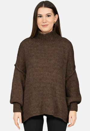 Woman wearing a loose brown knitted turtleneck sweater with long sleeves and black pants, standing against a plain background.