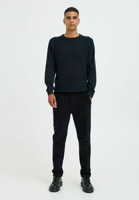 Tamno plavi crew-neck dukserica s teksturiranim materijalom, uparena s tamnim kordura hlačama i crnim čizmama na vezanje, stoji uz jednostavnu pozadinu.