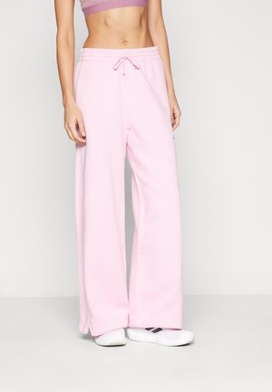 Femme portant un pantalon de jogging rose clair à jambes larges, des baskets blanches et un haut court mauve, debout devant un fond clair uni.