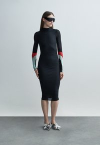 Nő áll, hosszú, fekete, testhez simuló ruhát visel, piros és világoskék ujjakkal, fekete napszemüveggel és ezüst hegyes orrú cipőkkel.